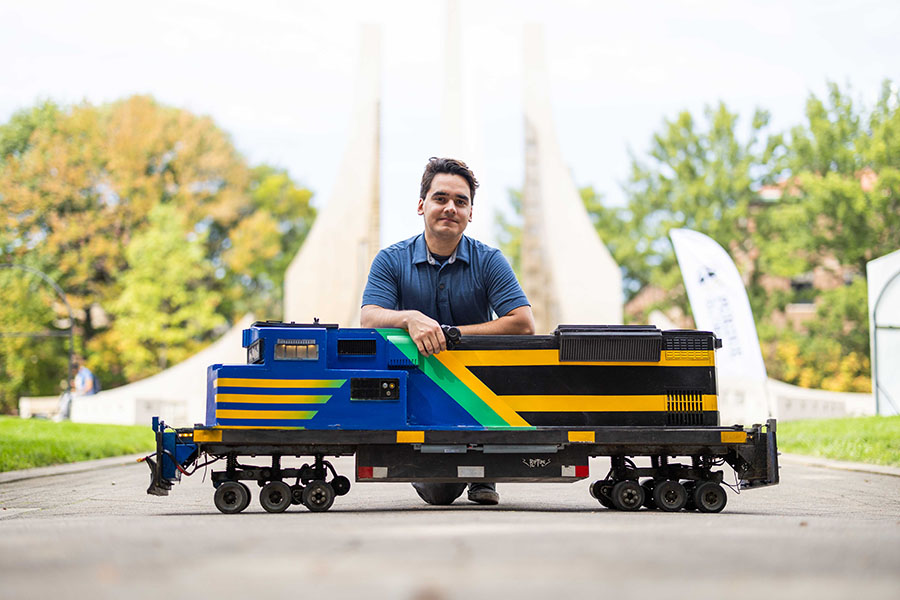 Student displaying locomotive skateboard, kneeling behind it, engineering fountain in background