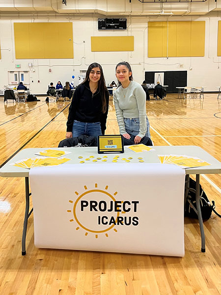 Two students standing behind a table with a tablecloth reading "Project Icarus"