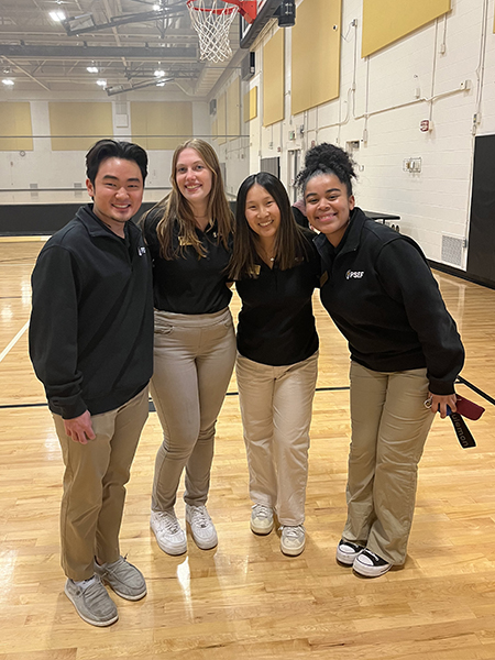 Four students inside the corec, smiling, wearing black polos