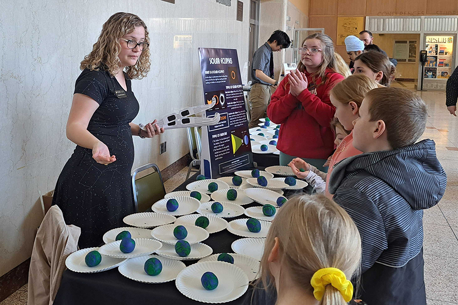 Woman with curly blonde hair wearing glasses standing behind a table with activities, speaking with students 