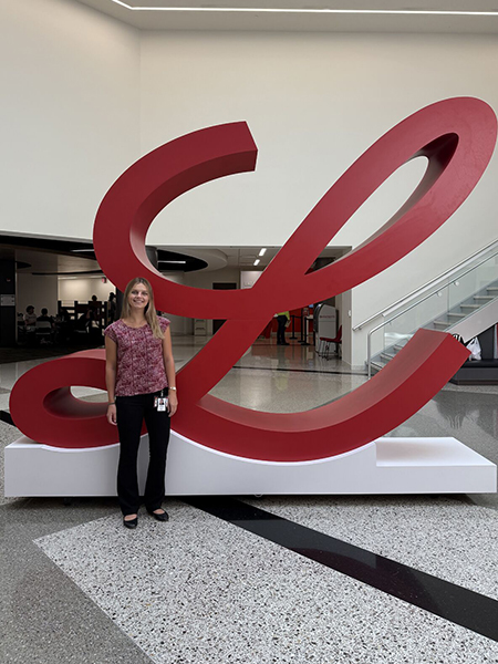 Woman with blond hair wearing pattern shirt and black pants standing front of a Lilly "L" structure