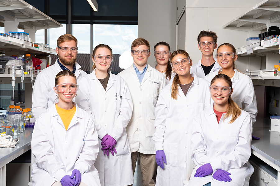 Group of students and a professor wearing lab coats, purple gloves and protective eyewear in a lab