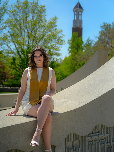 Student wearing white dress, gold stole, sitting near the engineering fountain