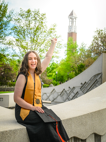Student wearing black dress, gold stole, dark brown hair, sitting near the engineering fountain