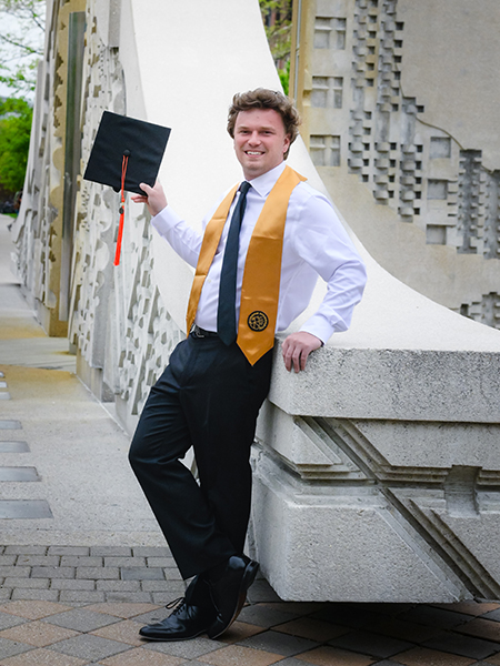 Student in black pants, white button-up shirt and black tie, gold stole and holding a graduation cap