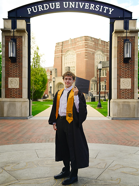 Student wearing black pants and white collared shirt, black graduation gown, gold stole and holding graduation cap, standing underneath Purdue arch