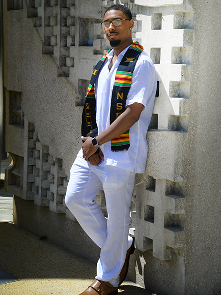 Student wearing white pants and white top, wearing NSBE stole, leaning against engineering fountain