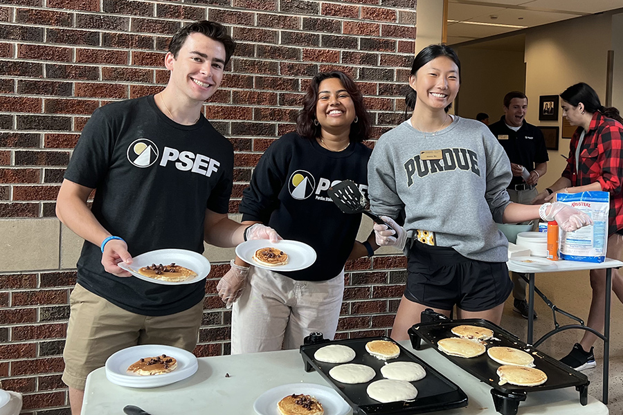 Three students smiling, standing behind a table with pancakes on grills