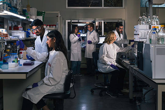 Purdue researchers collaborate with Lilly scientists in lab setting, wearing white lab coats and safety glasses.
