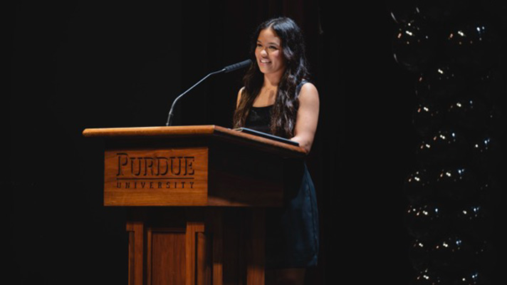 Woman with dark wavy hair, smiling, wearing black dress, standing behind lecturn