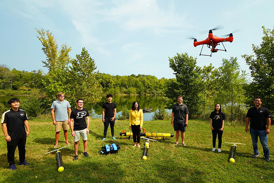 Group of people standing outside in front of a body of water watching a red drone fly in front of them.