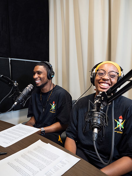 Two students wearing headphones, talking into microphone, laughing