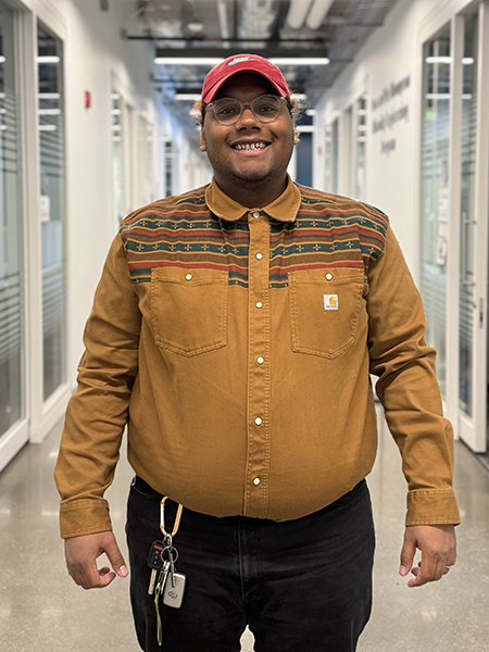 Student wearing glasses, a red hat and brown shirt, smiling, standing in hallway in Lambertus