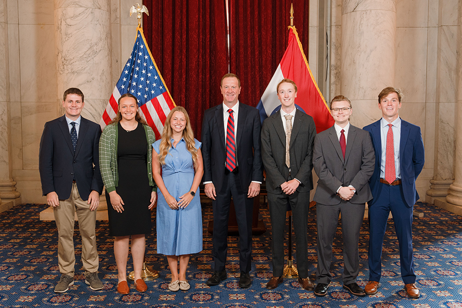 Group of students in business attire standing in front of a red curtain with two flags