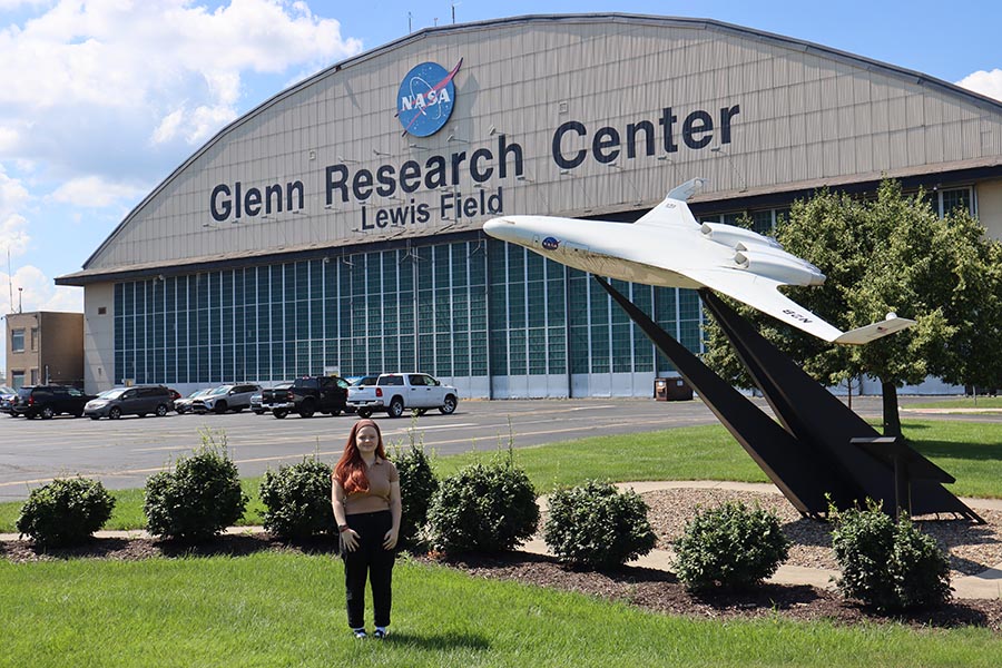 Woman stands next to airplane in front of brown hangar with blue NASA logo and black text that reads &ldquo;Glenn Research Center, Lewis Field.