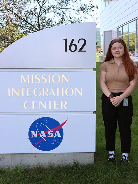 Woman stands next to white stone sign with blue NASA logo, black text that reads 162 and gold text that reads Mission Integration Center.