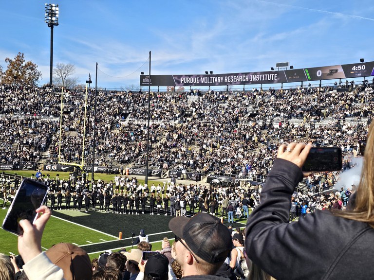 Zoomed out photo of many thousands of people in a stadium with a banner across the top back wall that reads "Purdue Military Research Institute".