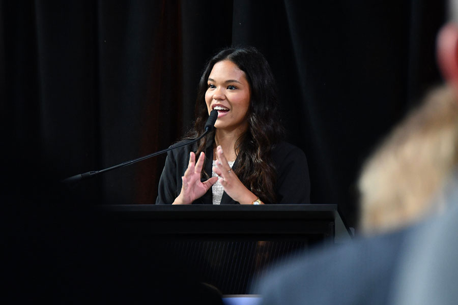 A student with dark hair and dark eyes, wearing a black blazer, speaking into a microphone at a press conference