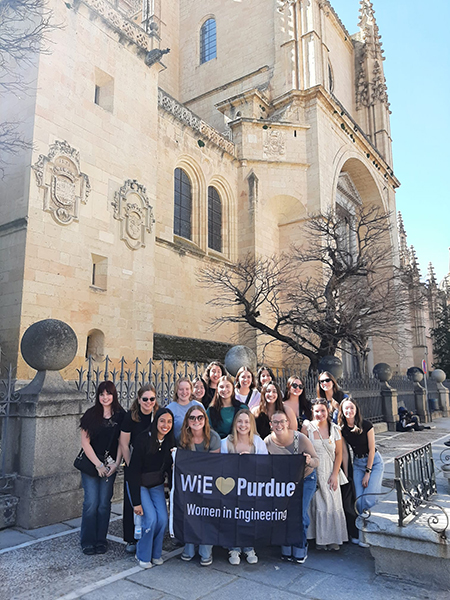 A group of 17 students, holding a banner that reads "WiE heart Purdue," stand in front of an old church during a study abroad trip